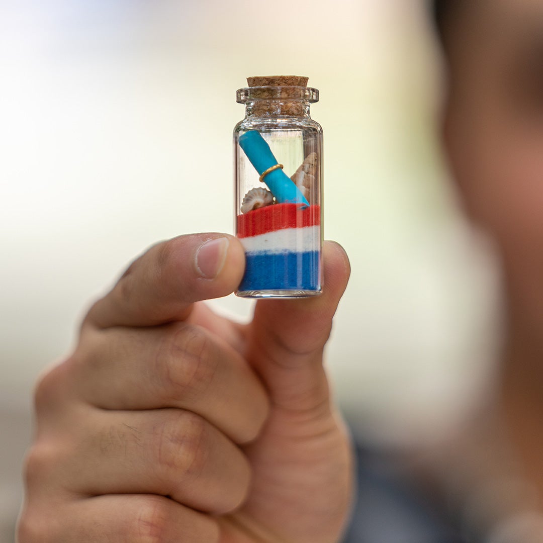 A student holds a tiny bottle with sand, a tiny shell, and a small note up to the camera.