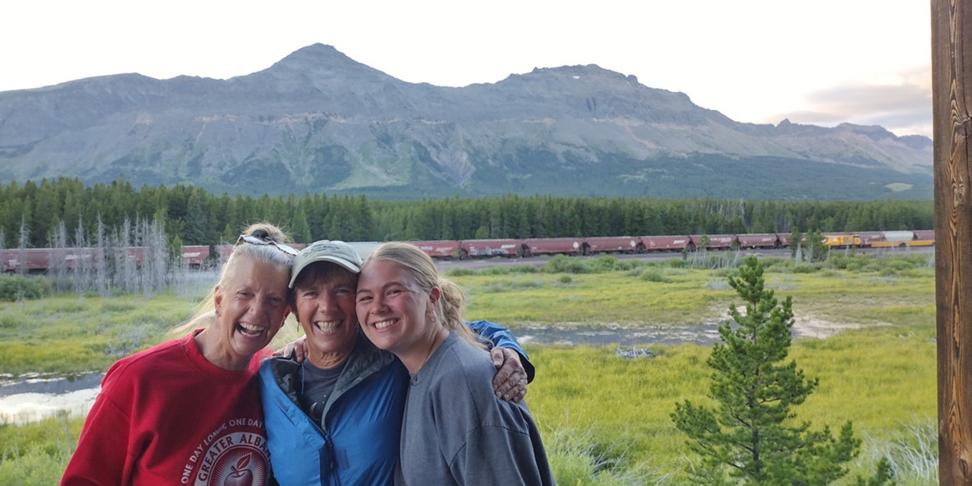 Three women of varying ages hugging in front of a vast landscape with grassy plains and a mountain in the distance