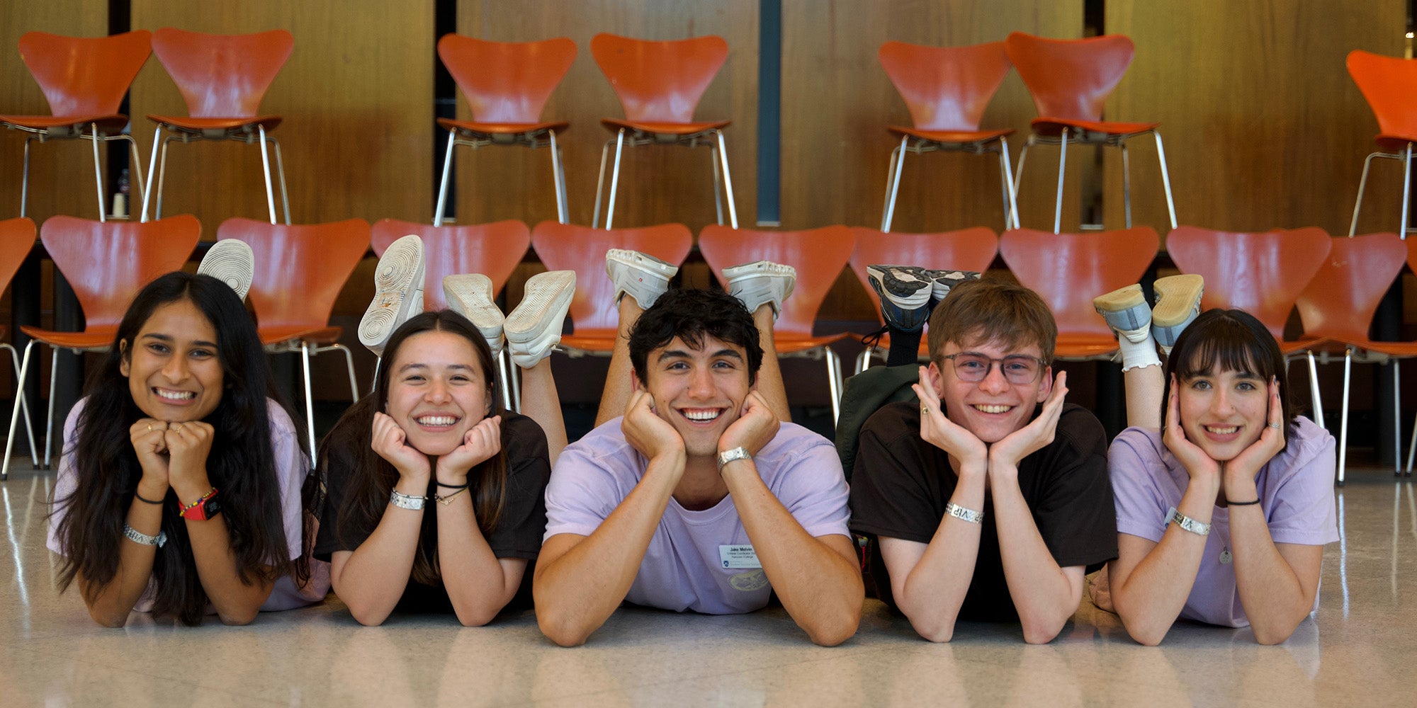 Five people lying on their bellies on the floor in identical poses, propped up on their elbows, holding their heads up with their hands, legs up behind them