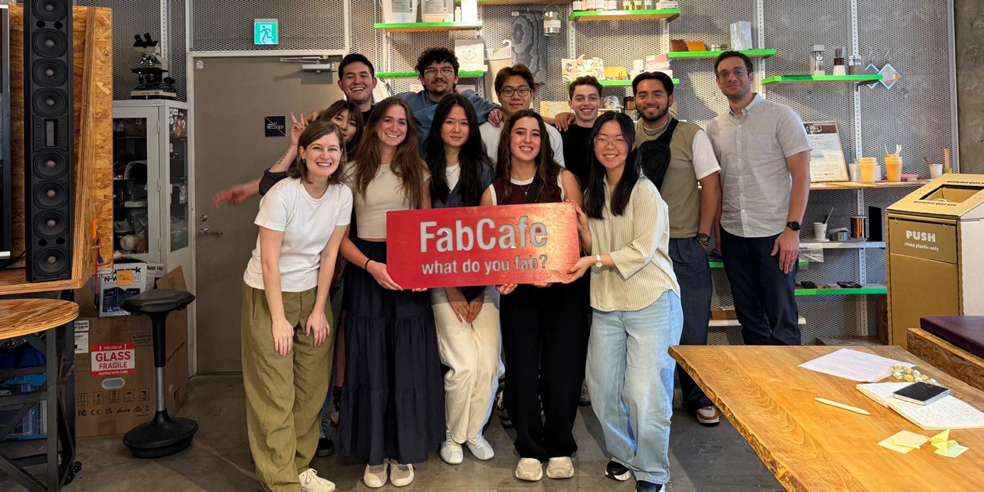 Large group of people holding a banner that says "FabCafe" in a workshop