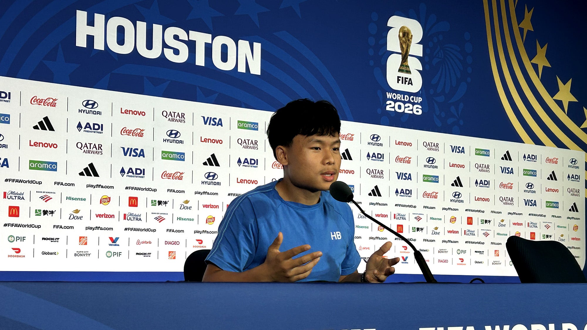 A guy sitting in a broadcasting booth, with several sponsor and FIFA World Cup logos behind him