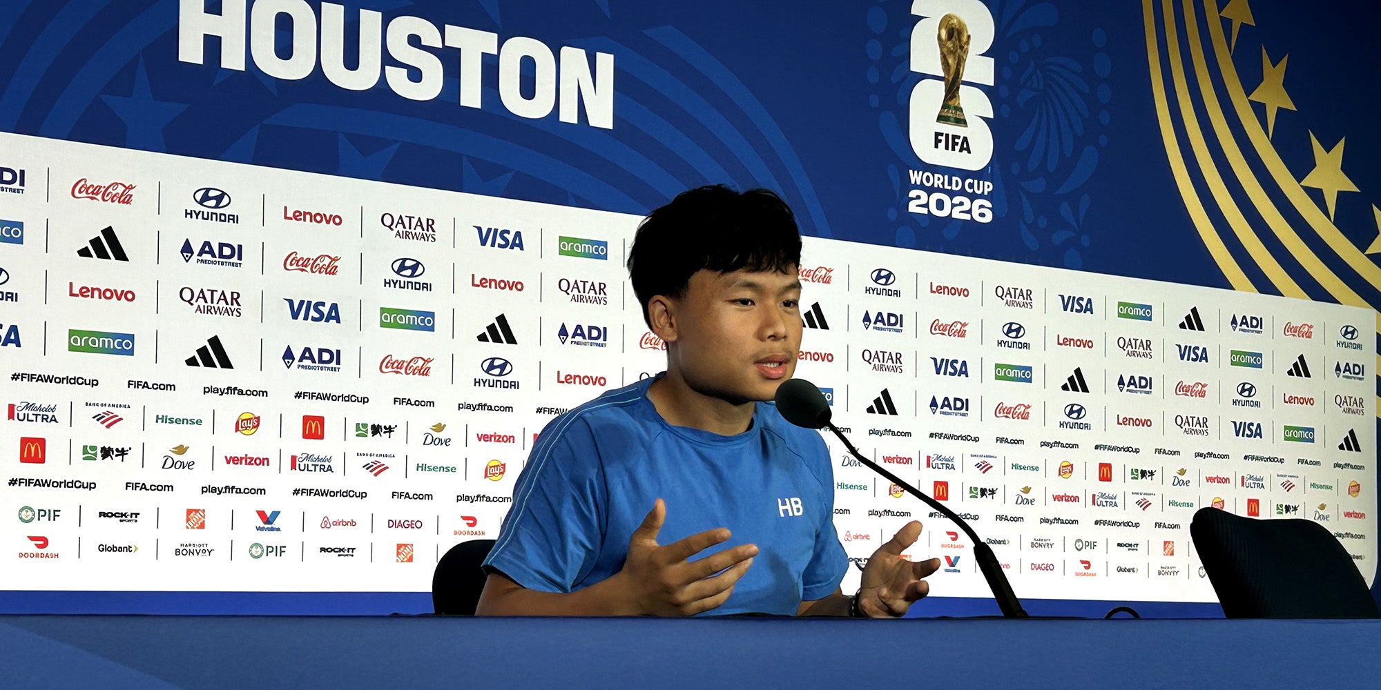 A guy sitting in a broadcasting booth, with several sponsor and FIFA World Cup logos behind him