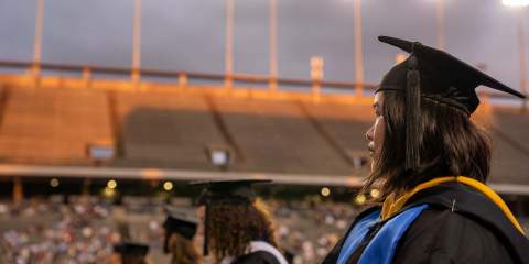 A female student wearing her graduation cap and gown is in the foreground looking to the side at the graduation ceremony being held in Rice Stadium.