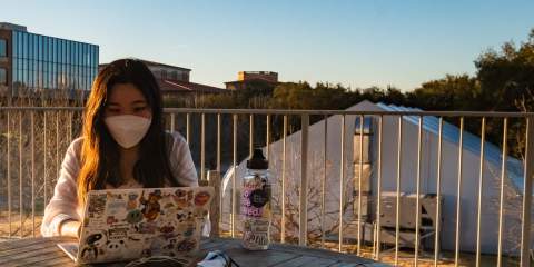 A girl wearing a mask sits at a table outside at sunset on Rice University's campus with her laptop open in front of her.
