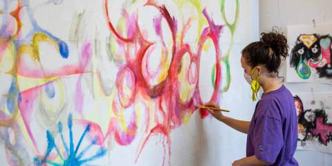 A female student wearing a purple shirt paints on a wall of red, yellow and green overlapping circles, with a watercolor tinge to the paint.
