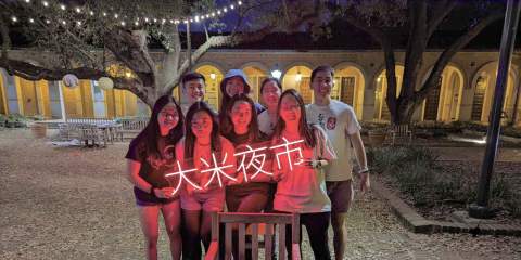 A group of male and female students from the Rice Taiwanese Association smile and pose with a neon sign that says "Rice night market" in Mandarin.
