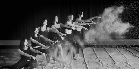A black and white photo of an all female group of six contemporary dancers pose in a line with their hands out, after throwing chalk which makes a cloud to the right of them.