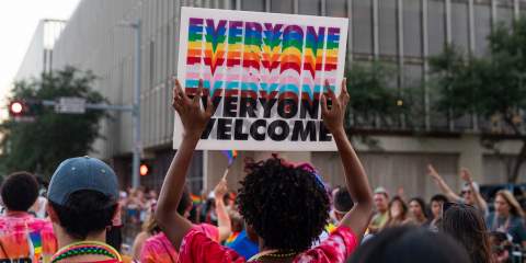 This photo is taken from behind a Rice participant in the Houston Pride Parade holding a sign above their head that says Everyone Welcome in the colors of the pride flag.