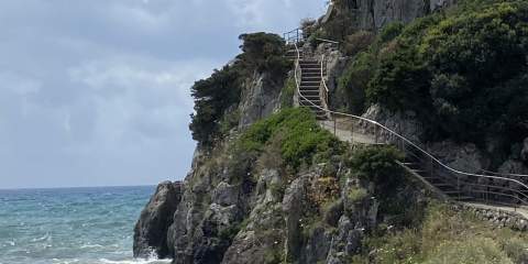 A picture of a cliff and the water below in Cosa, Italy, as well as a staircase that goes up the side of the cliff in the right side of the picture.