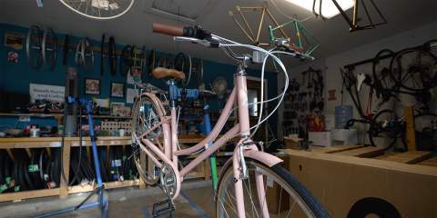 Inside of a garage shop with wheels and gears hoisted along the walls and a pink bike in the center of the frame.