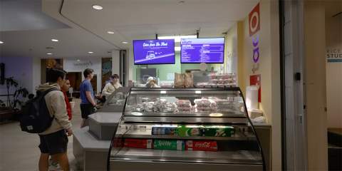 Wide view of clear food display case with food inside and two tv screens above the case. Students are lined up to buy food.
