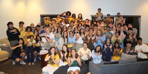 A group of college students, many in yellow t-shirts, posing in a dorm hall and making owl wings by connecting their hands at the thumb