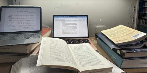 A pile of textbooks and papers stacked around a laptop on top of a desk