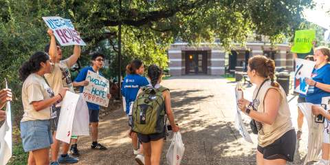 A student with a backpack walks follows an Owl Guide toward a building, being cheered on by fellow students with "welcome" posters