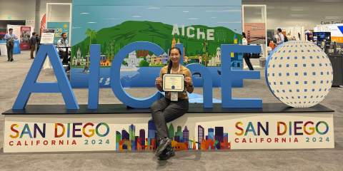 A girl holding a certificate in front of large display letters that say "AIChE"