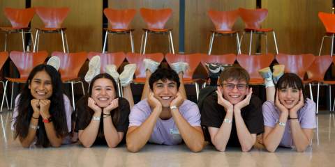 Five people lying on their bellies on the floor in identical poses, propped up on their elbows, holding their heads up with their hands, legs up behind them