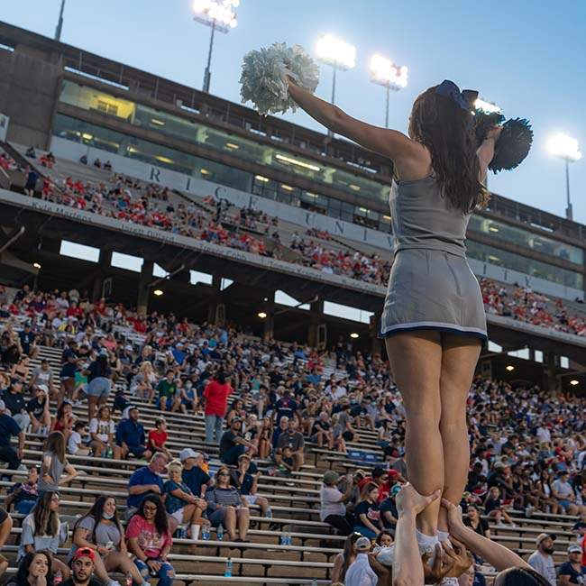 A female cheerleader is pictured from the back holding a white pom pom and a navy pom pom. She is being held up with her hands in a V to cheer on the crowd in front of her.