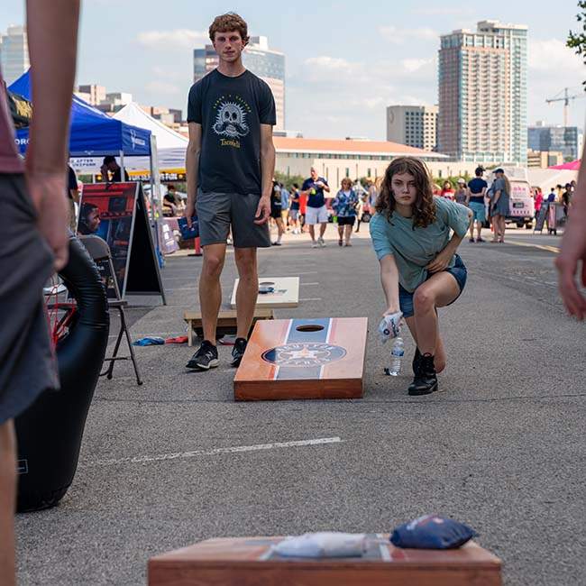 Two students play cornhole, with the student on the right bending down getting ready to toss the bean bag in the opposite board.