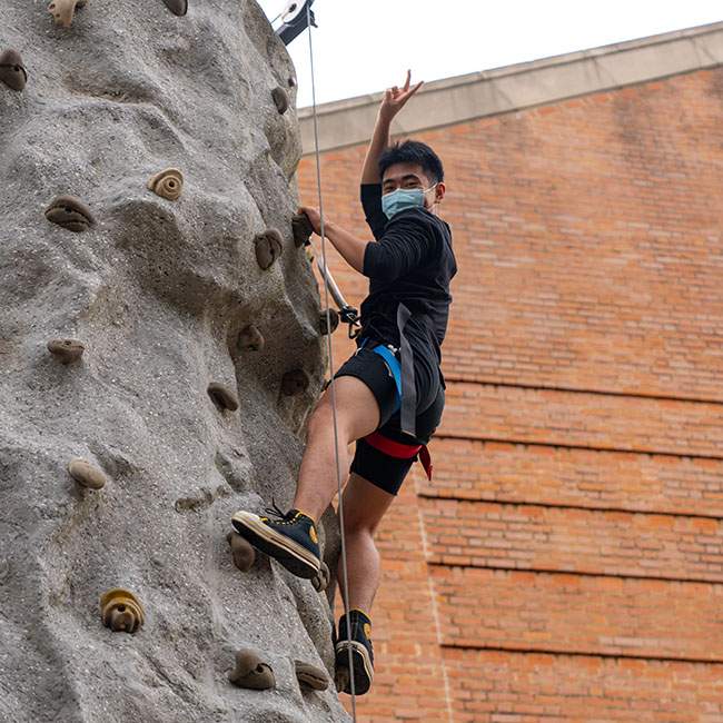 A male Rice student climbing a rock wall raises one arm making a peace sign with his fingers as he nears the top.