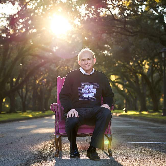 President Leebron sits in a chair on Rice's campus wearing a sweatshirt designed by student club Rice Design.
