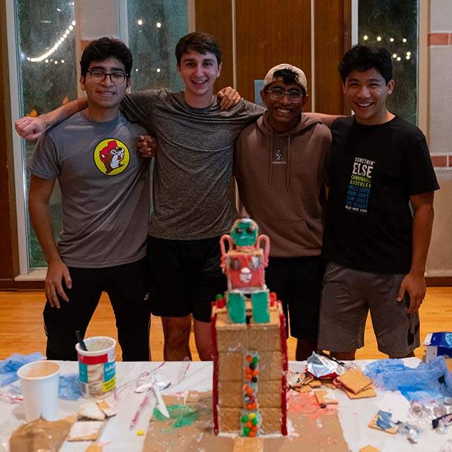 A group of four male students poses with their gingerbread house.