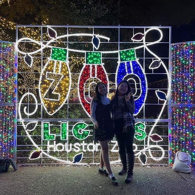Grace, a Rice student, poses with Sarah, a Rice alumna, in front of a lit-up sign that says "Zoo Lights Houston Zoo."