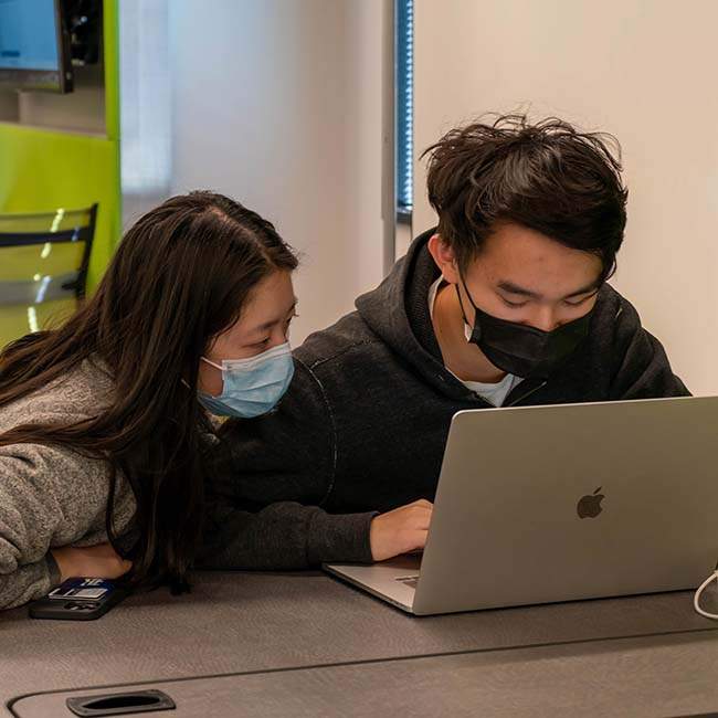 A female and a male student both wearing masks look at the laptop that the male student is typing on.