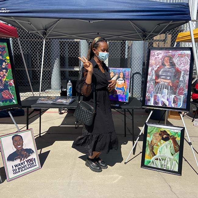 A female student poses in front of a booth filled with framed art showcasing Black excellence and the Black experience.