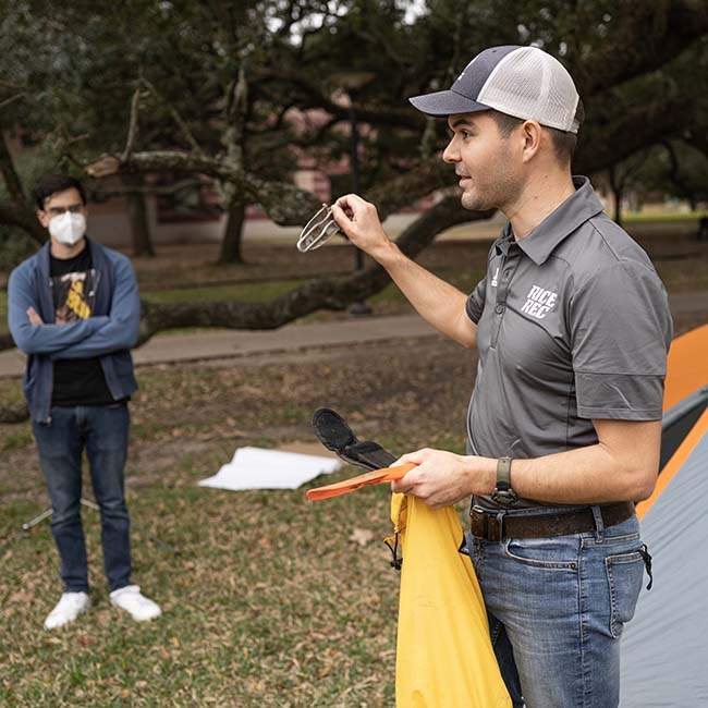 A man holding a piece of a tent gestures while giving a talk on camping, with a student standing off to the side listening.