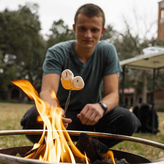 A male Rice student holds a stick with two marshmallows over a small flame.