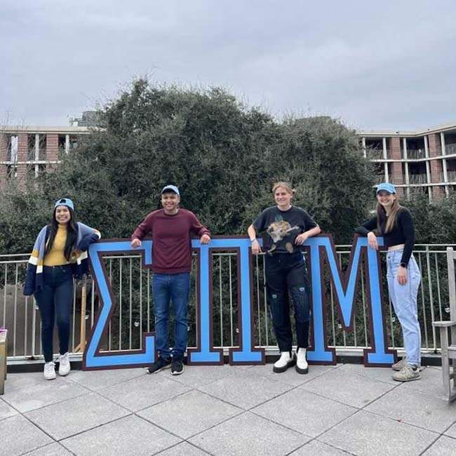 Four students stand with large Greek letters that represent Martel - Sigma Pi Mu.
