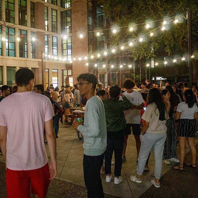 A large group of students gather around an outdoor patio at nighttime mixing and mingling with a string of lights overhead.
