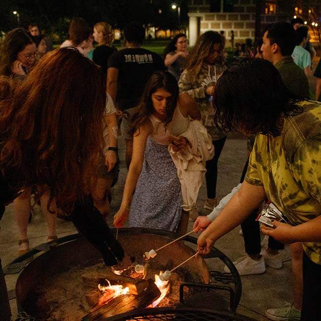 Three students hold out sticks with marshmallows on them over a grill to make s'mores.