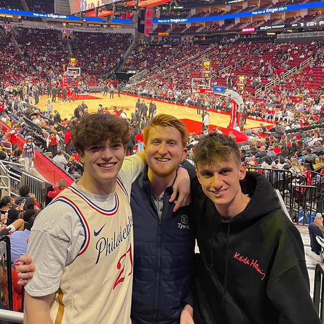 Three male Rice students smile and pose for a picture in the stands of a Houston Rockets game, with the basketball court behind them.