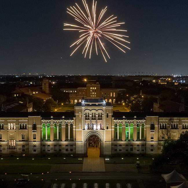 A drone picture of Lovett Hall at nighttime shows the building lit up and a single firework exploding above.