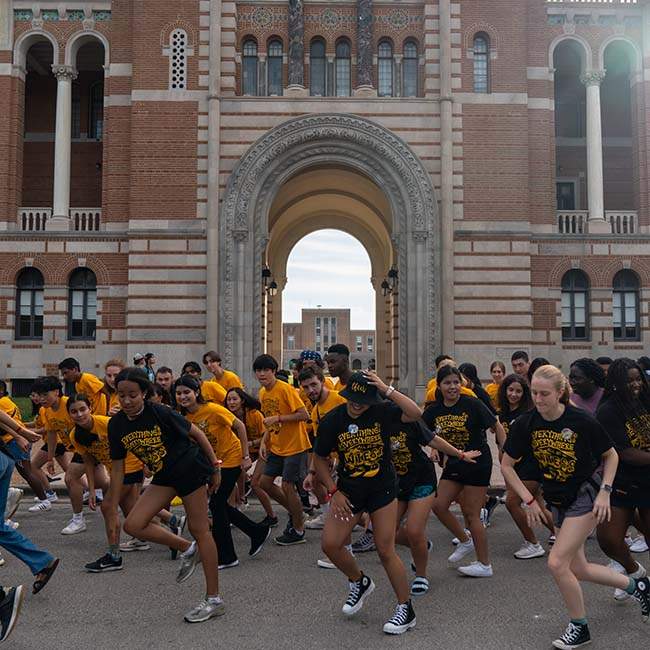 A large group of students from Wiess Residential College wearing their college colors, goldenrod and black, perform their O-Week Dance in front of the Sallyport of Lovett Hall.