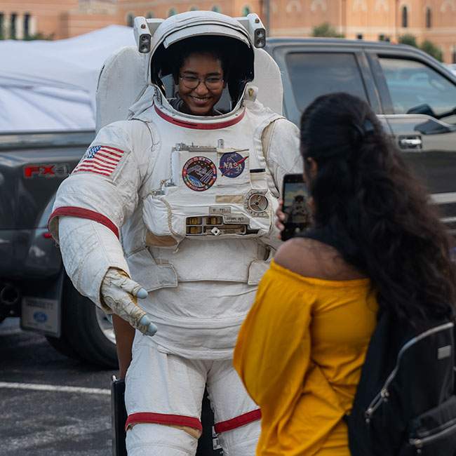 The picture shows the back of one student taking a picture of another student in front of her that looks like they are wearing a space suit.