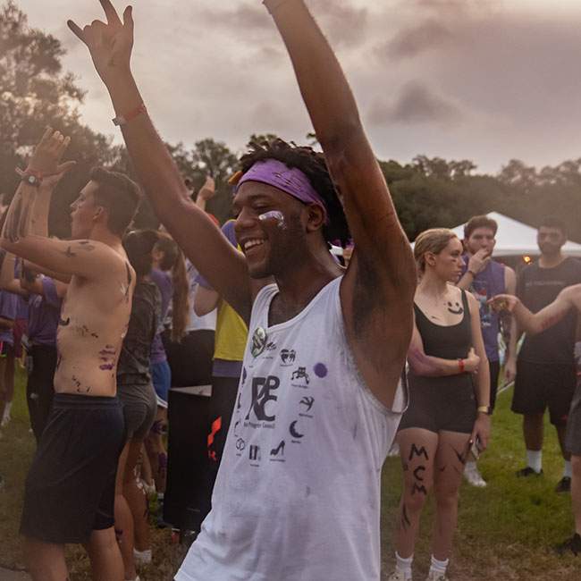 A Rice student with purple body paint for McMurtry College holds both hands up in the air while smiling in the middle of the crowd of new students at an O-Week event.