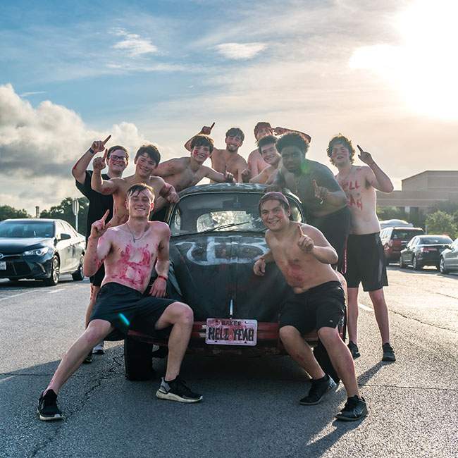 A group of Baker college boys covered in red body paint for their college color smiles for a group photo while leaning against an old Volkswagen Beetle Car that is decorated with Baker College’s symbol. 
