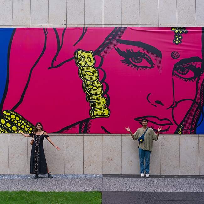 Devika and Naman stand in front of a large piece of art that is of a woman in bright pink drawn in the pop art style.