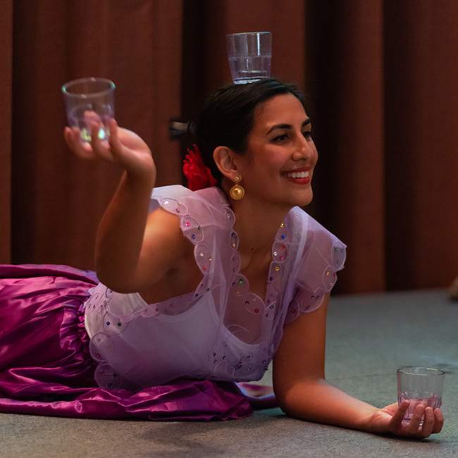 A woman dressed in traditional Filipino clothing is doing a dance where she lays on the ground and is holding two glass cups in her hands and balancing one glass cup on her head.