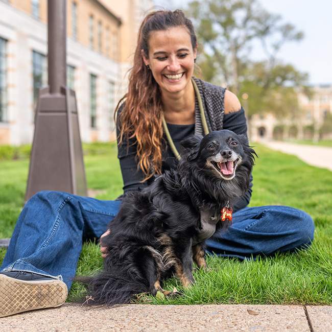 A student sitting on the ground outside with a small dark brown dog in her lap