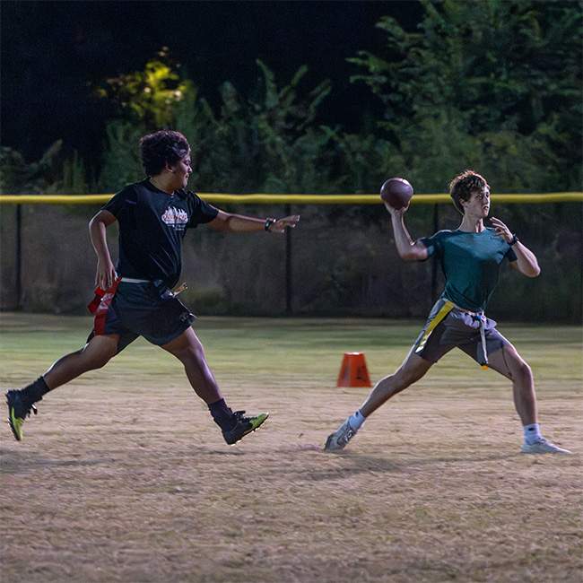 A boys prepares to launch a football outdoors at night, while another chases after him.