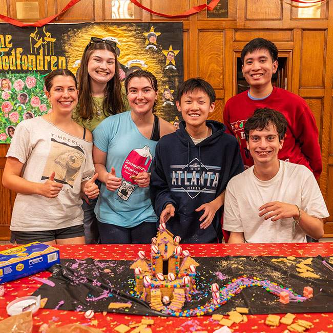 Students pose behind a table with a graham-cracker house sitting on it