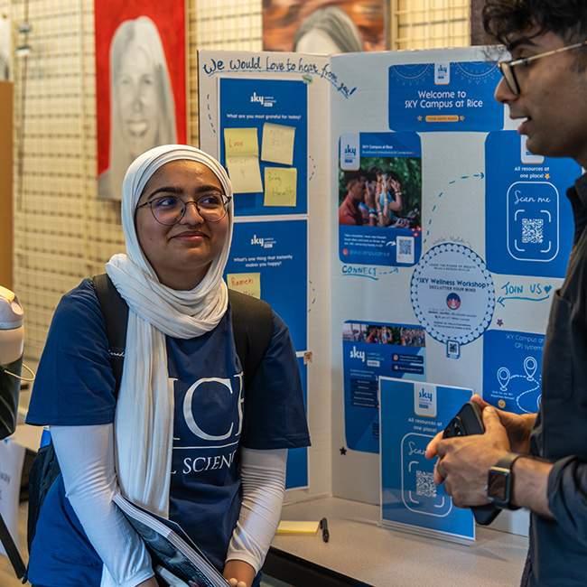 A girl in front of a poster board, looking up at a guy speaking partly out-of-frame