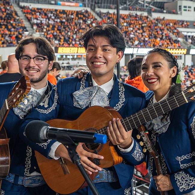 Three people pose for a photo, the middle man is hugging a guitar, crowd visible behind them