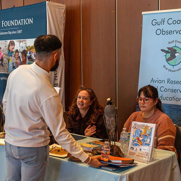 Two women sitting behind a table; a man standing in front of it