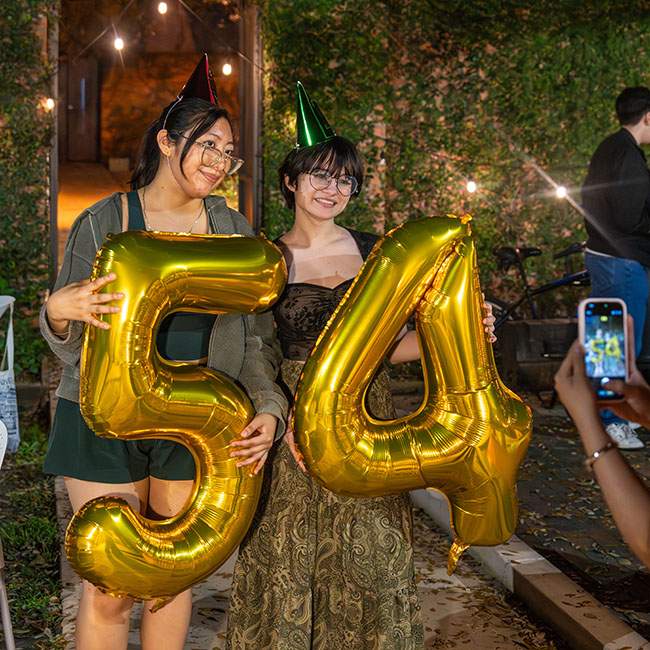 Two girls hold up gold inflatable balloons in shape of "5" and "4"