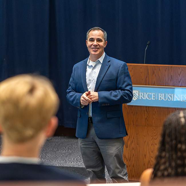A man in a blue suit standing on a stage, speaking to partially visible students in the foreground