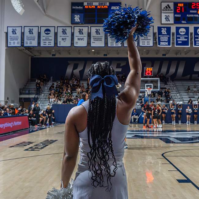 Back of a cheerleader lifting a pom pom in the air, facing the court & seating area